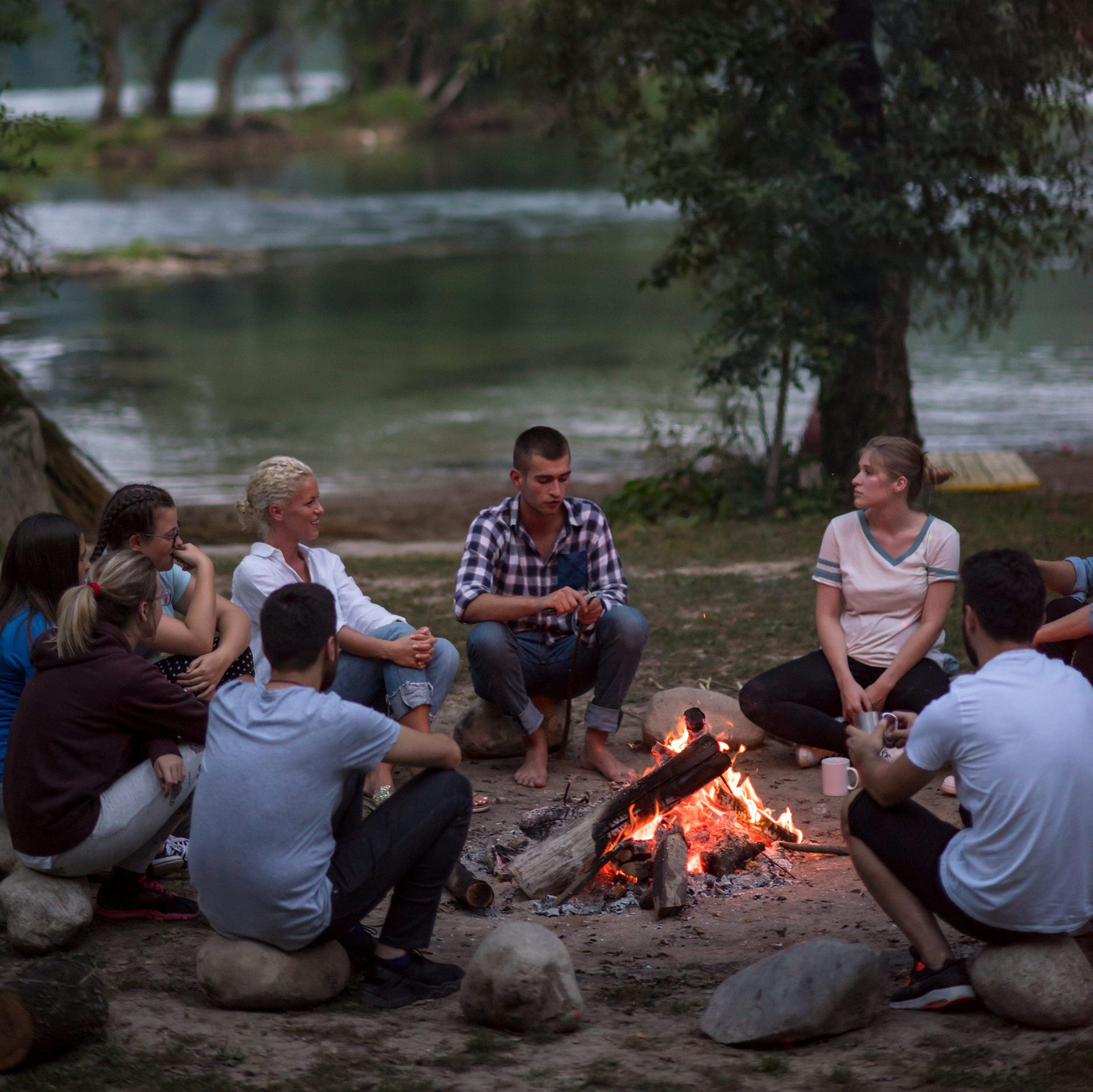 a group of happy young friends relaxing and enjoying summer evening around campfire on the river bank